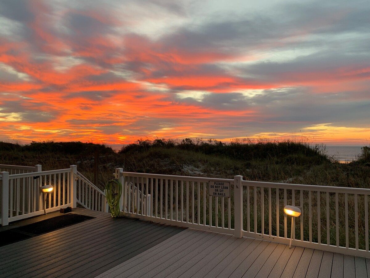 A wooden deck with railings and a warning sign, overlooking a beach at sunset.