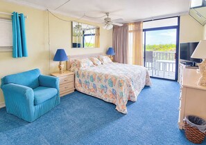 A hotel room with a bed, a chair, a desk with a lamp, a television, and a balcony with a view.