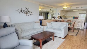 Un salon avec un canapé, un fauteuil et une table basse, un coin repas avec des chaises, et une cuisine à l’arrière-plan.