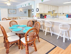 A dining area with a glass table surrounded by wicker chairs, a kitchen with white cabinets, and a small island with stools.