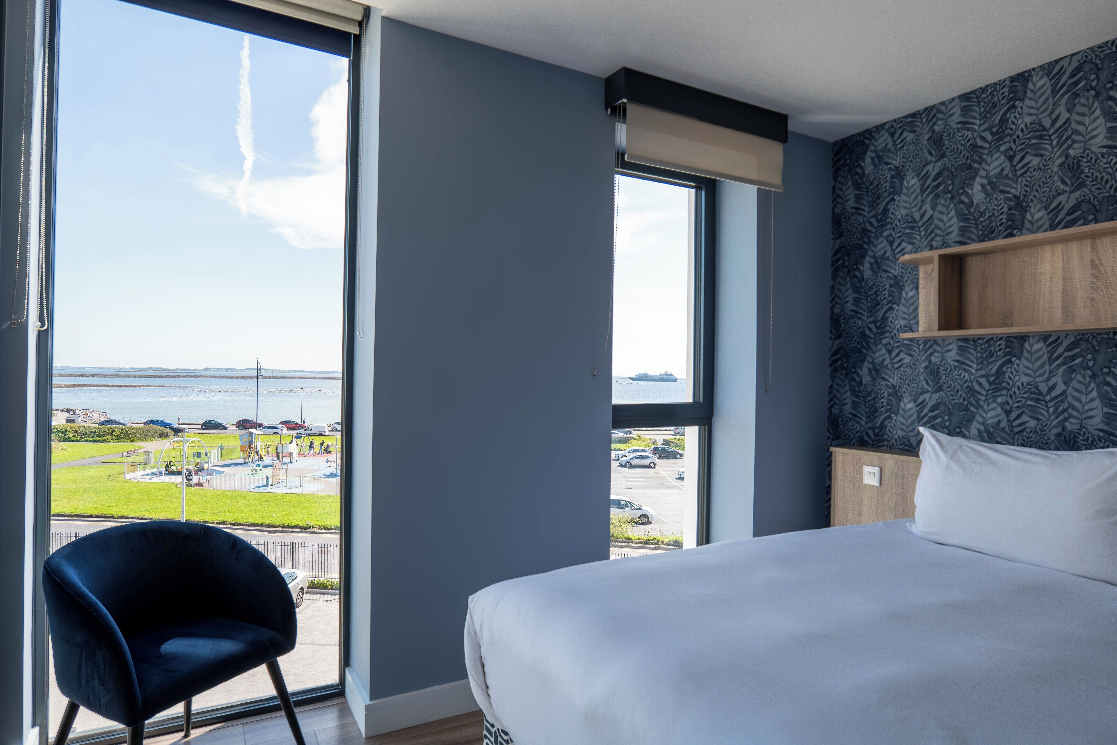 Une chambre d’hôtel dotée d’une grande fenêtre donnant sur un parking et un plan d’eau. On y trouve un lit avec une literie blanche et un fauteuil bleu foncé.