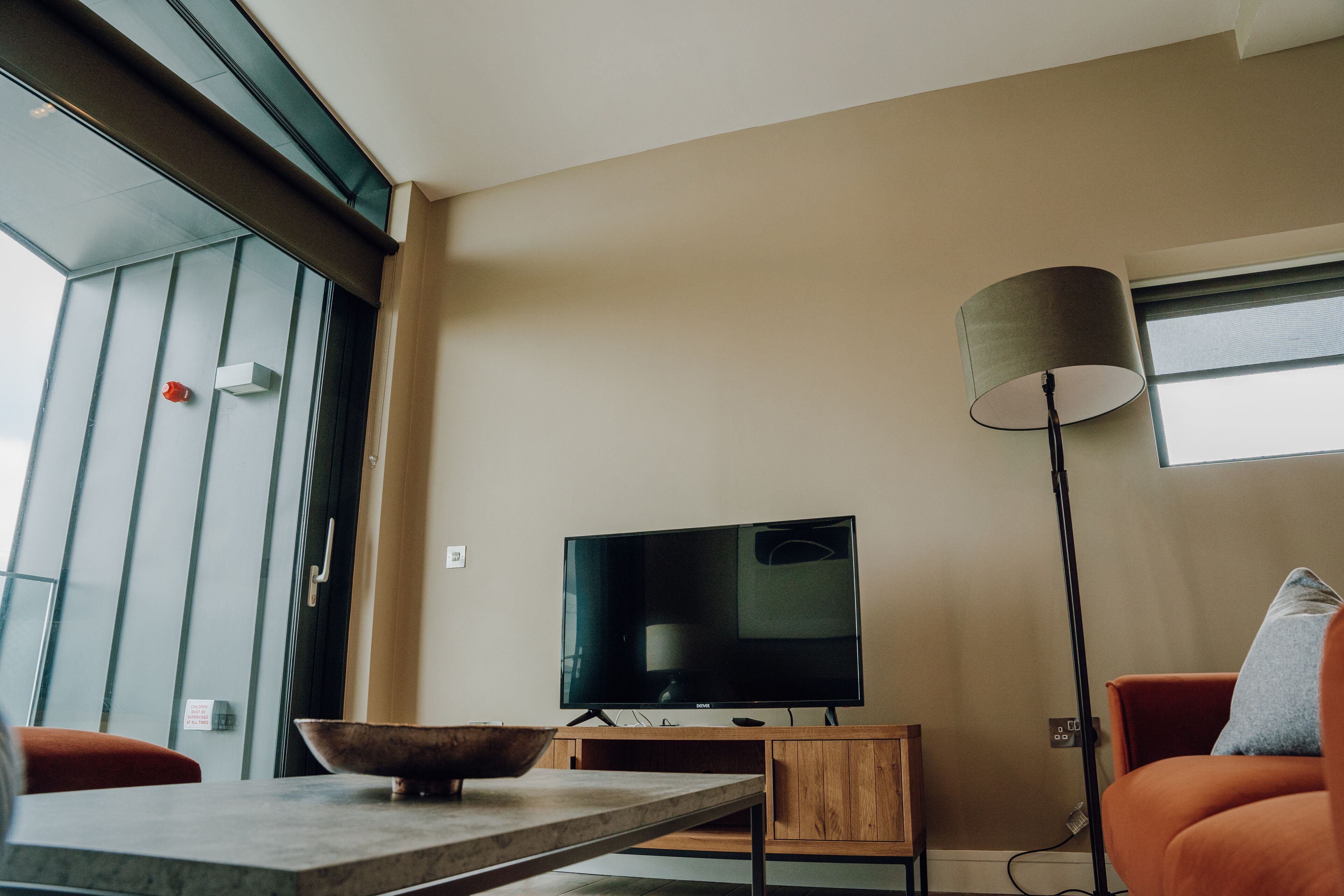 A modern living room with a flat-screen TV on a wooden stand, a floor lamp, and a dining table with a bowl.