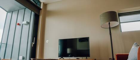 A modern living room with a flat-screen TV on a wooden stand, a floor lamp, and a dining table with a bowl.