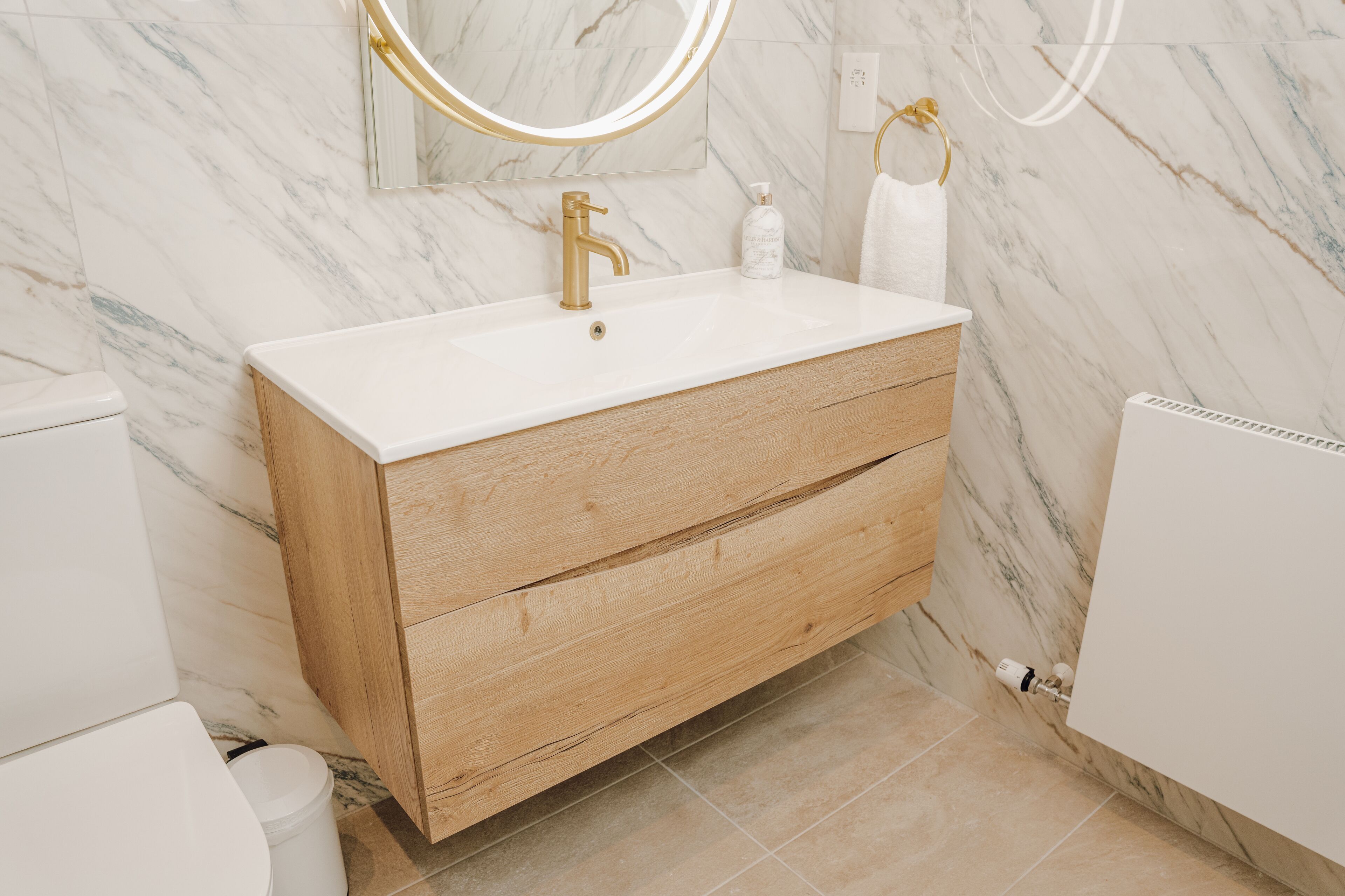 Une salle de bain moderne avec un mur en marbre, un meuble-lavabo en bois, un robinet doré et un miroir rond.