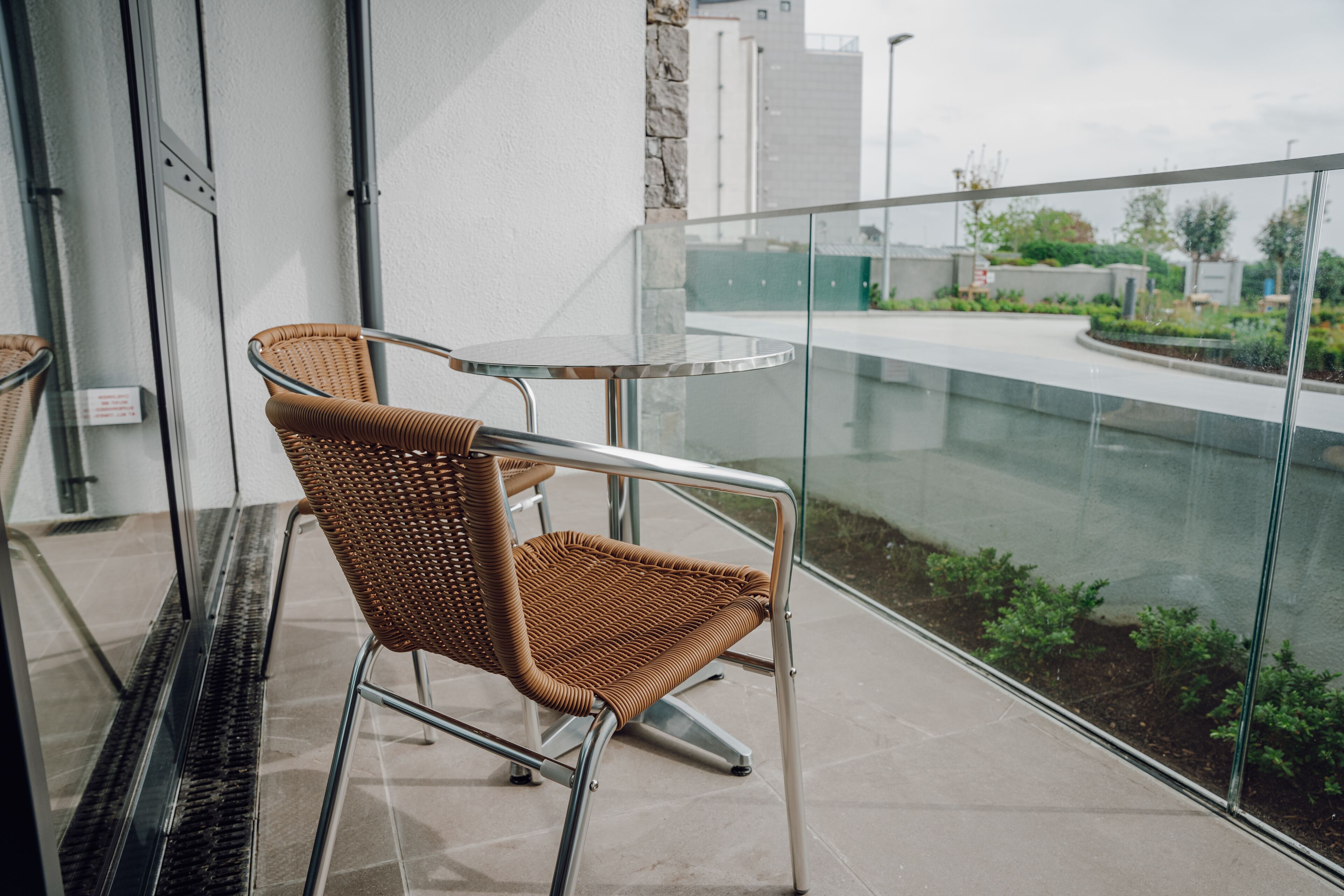 A wicker chair with a metal frame on a balcony with a glass railing, overlooking a landscaped area with greenery and buildings in the background.