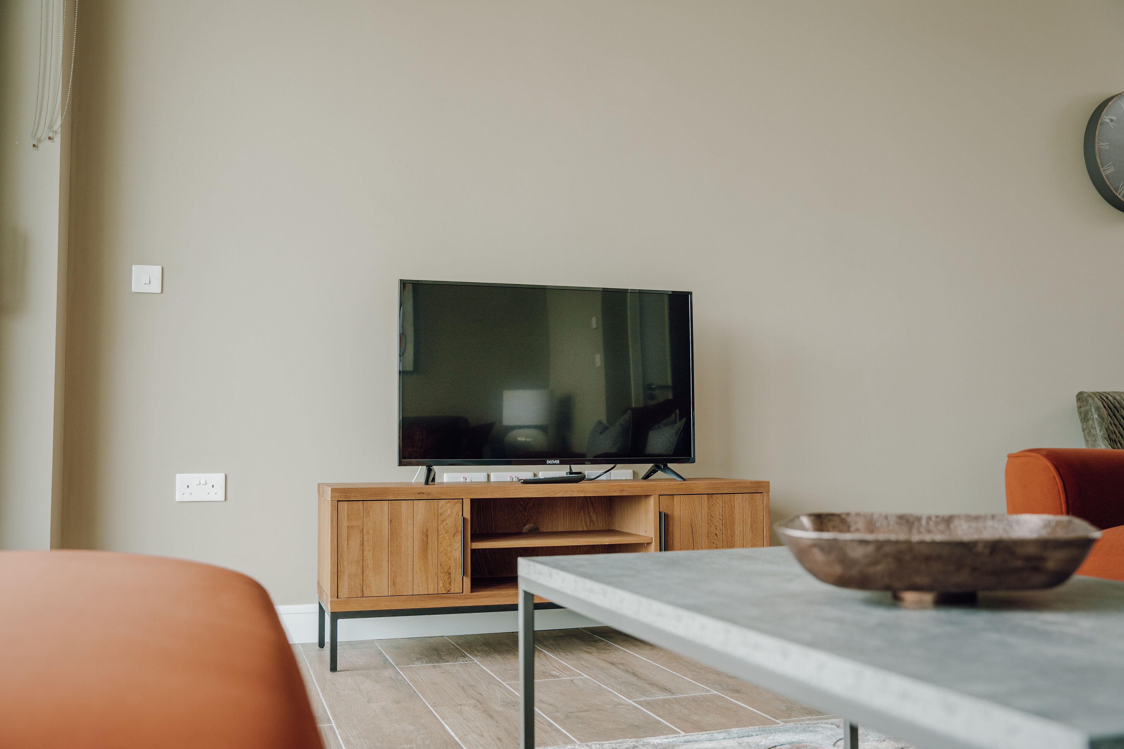 A modern living room with a flat-screen TV on a wooden stand, a dining table with a bowl, and orange chairs.