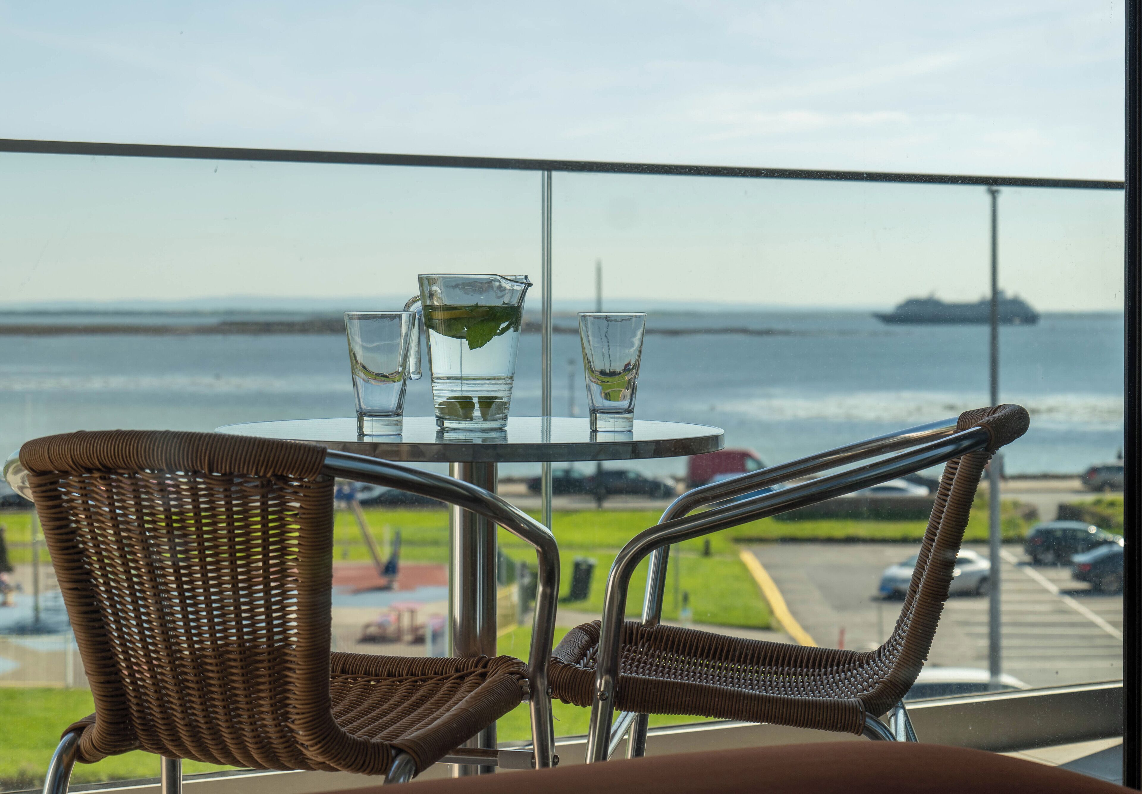 A balcony with wicker chairs and a table, overlooking a body of water and a distant ship.