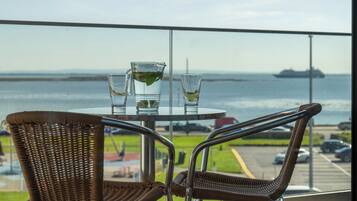 A balcony with wicker chairs and a table, overlooking a body of water and a distant ship.