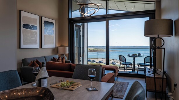 A modern living room with a view of the sea, a dining area with a table set for two, and a large window. - Shoreline Apartments (Galway)
