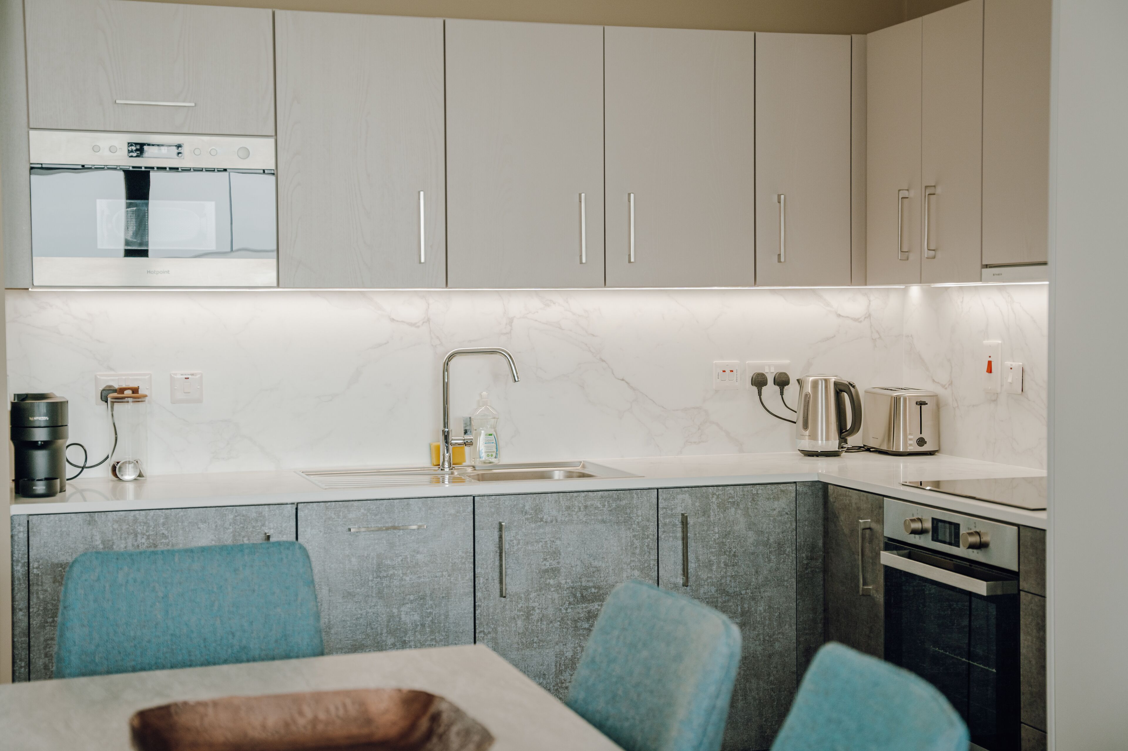 A modern kitchen with grey cabinets, a white marble backsplash, a built-in oven, a coffee maker, and a kettle.