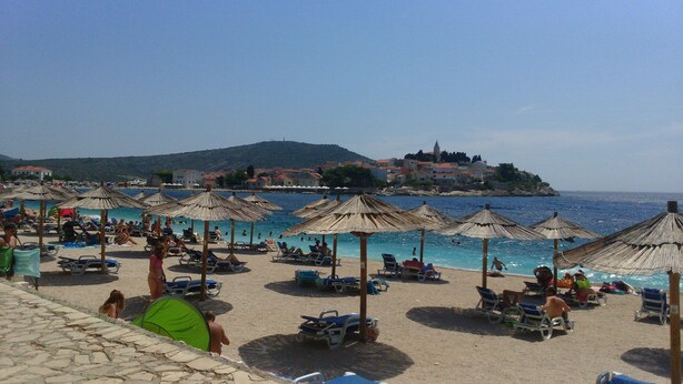 A beach with umbrellas and lounge chairs, a clear sky, and buildings in the background.
