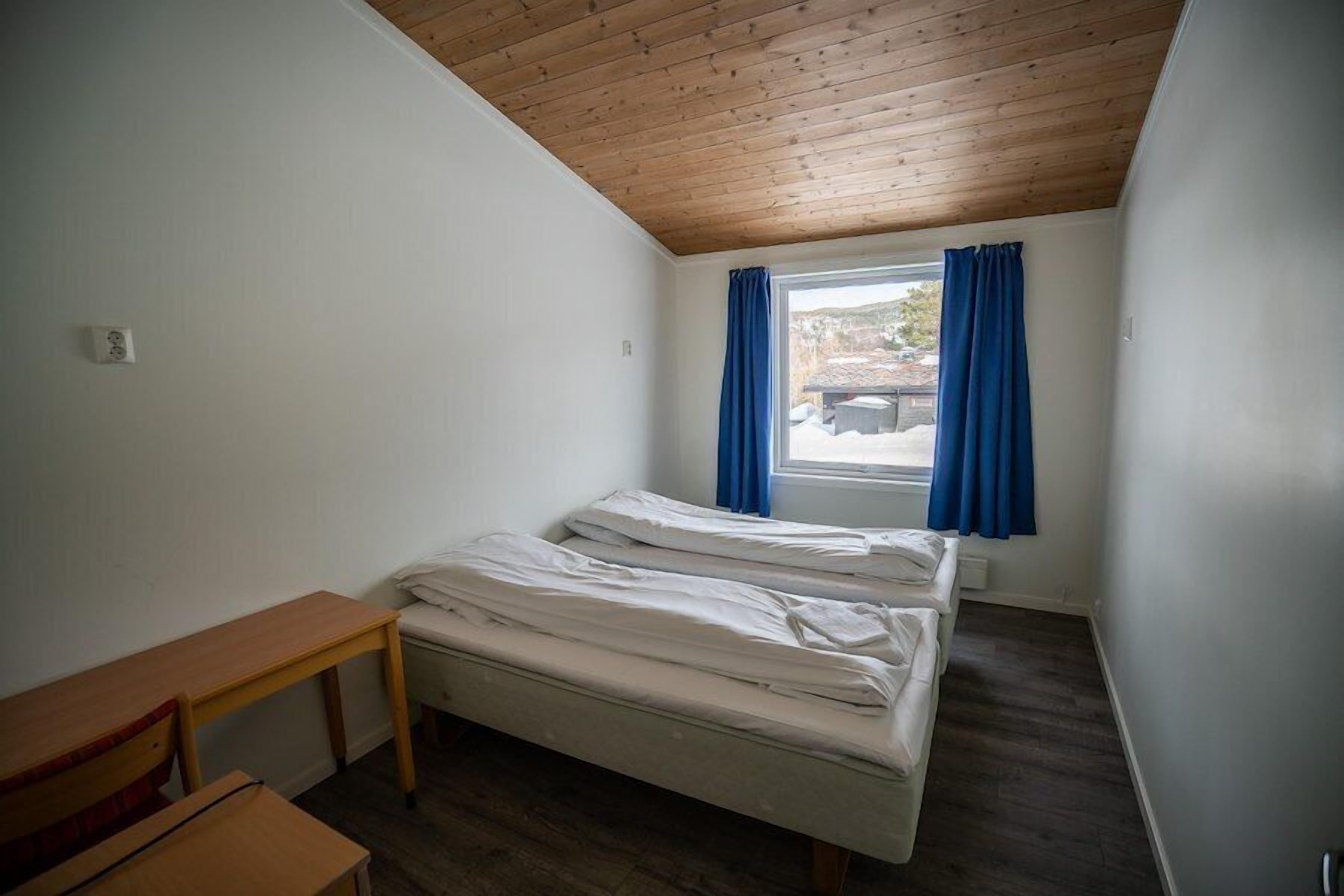 A small, simple room with two beds, a wooden desk, and a window with blue curtains.