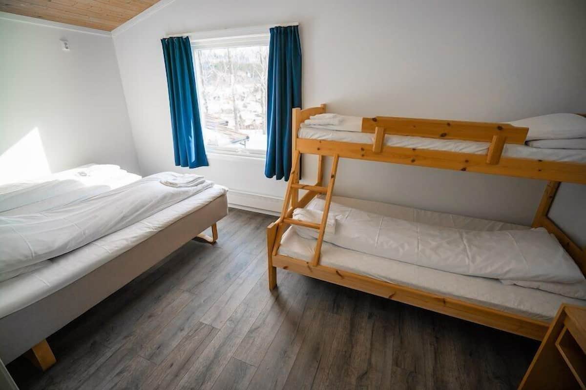 A room with two bunk beds, a window with blue curtains, and a wooden floor.