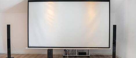 Una pantalla de proyección blanca de gran tamaño con marco negro, flanqueada por dos altavoces negros a cada lado, montada sobre una pared blanca con suelo de madera.