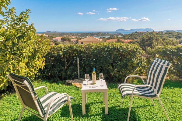 Une table avec deux chaises et une bouteille de vin, placée sur une pelouse d’où l’on peut admirer un vignoble et des maisons au loin.
