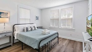A bedroom with a bed, nightstands, a desk, and two windows with blinds. - Orange Door Bungalow - Central, Cozy, 3BR (Jacksonville)