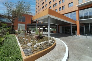 Un edificio moderno con un acceso cubierto, un jardín ajardinado con un estanque y un cielo despejado.