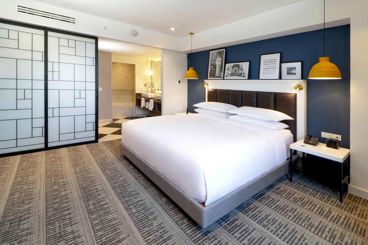Habitación de hotel con una cama grande, mesita de noche con teléfono y lámpara, y un baño visible a través de una puerta abierta, además de una alfombra estampada.