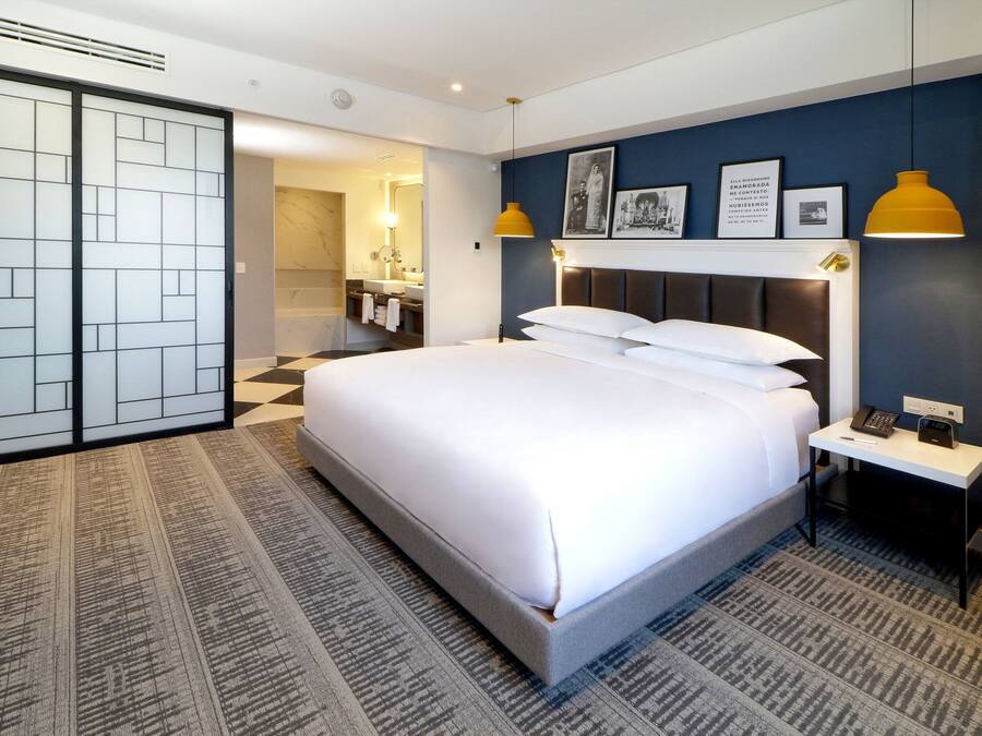 Habitación de hotel con una cama grande, mesita de noche con teléfono y lámpara, y un baño visible a través de una puerta abierta, además de una alfombra estampada.