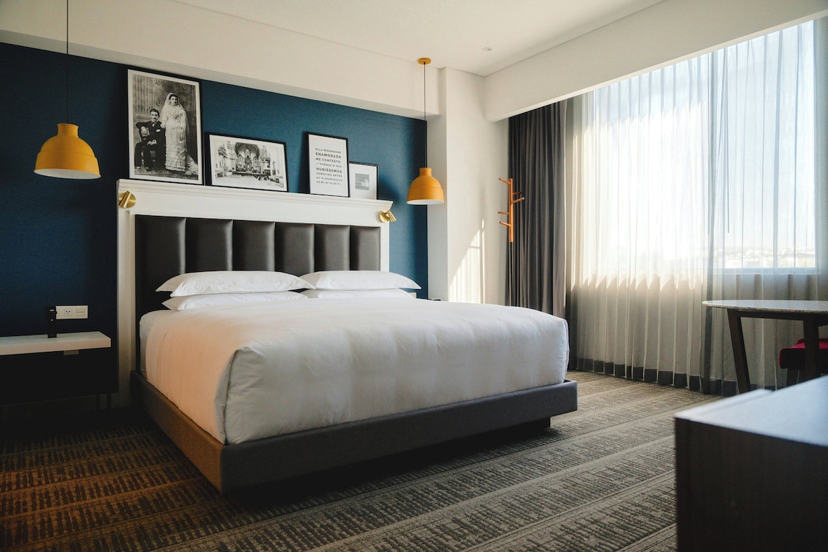 Habitación de hotel con una cama grande, cabecera con panel de color claro, mesita de noche pequeña, escritorio con silla, ventana grande con cortinas y dos lámparas colgantes.