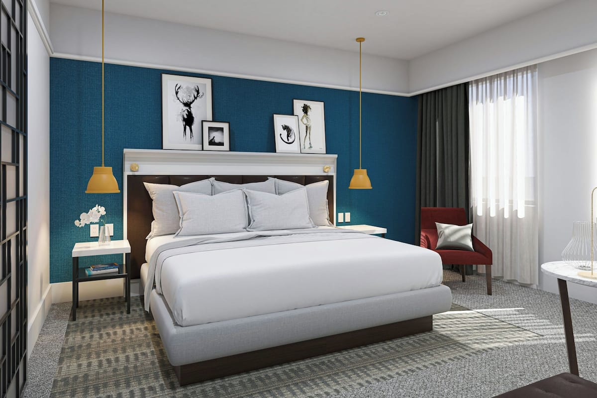 Una habitación de hotel moderna con una cama grande, un sillón rojo, una mesita pequeña y obras de arte en la pared.