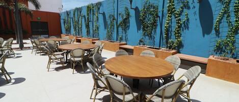 An outdoor patio with round wooden tables and wicker chairs, surrounded by a blue wall with climbing plants.