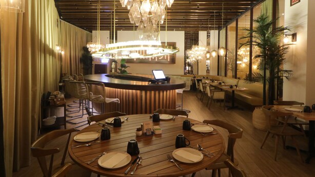 Un restaurante con mesas de madera preparadas para comer, una zona de bar al fondo y un candelabro colgando del techo.