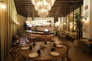 Un restaurante con mesas de madera preparadas para comer, una zona de bar al fondo y un candelabro colgando del techo.