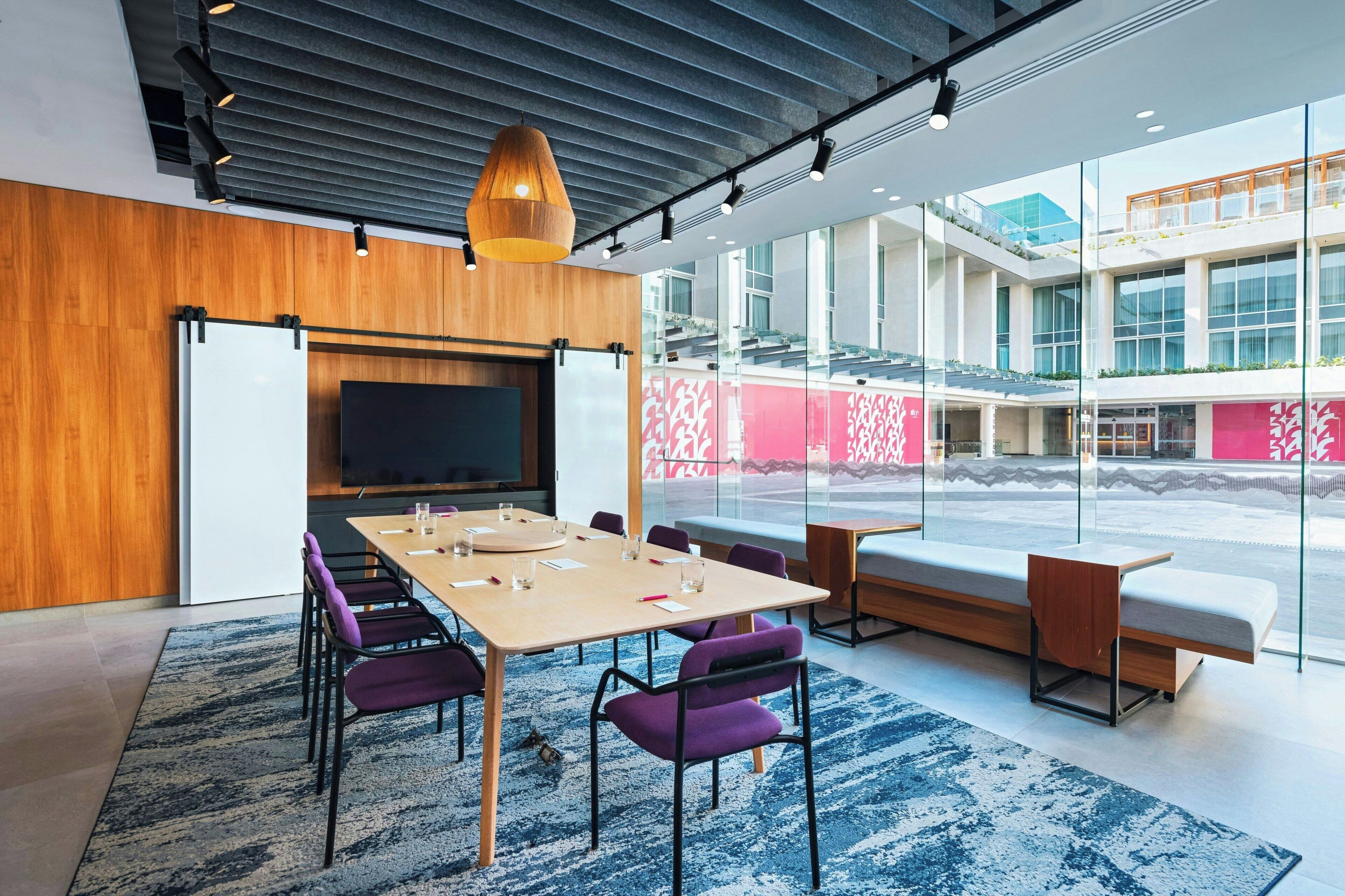 Een moderne vergaderruimte met een grote tafel, paarse stoelen, een televisie en een groot raam met uitzicht op een gebouw.
