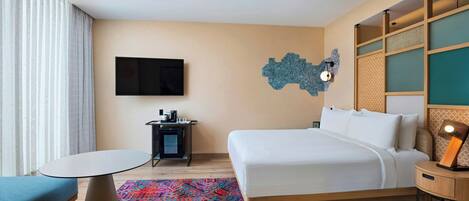 A hotel room with a bed, a desk, a chair, a TV, and a patterned rug.