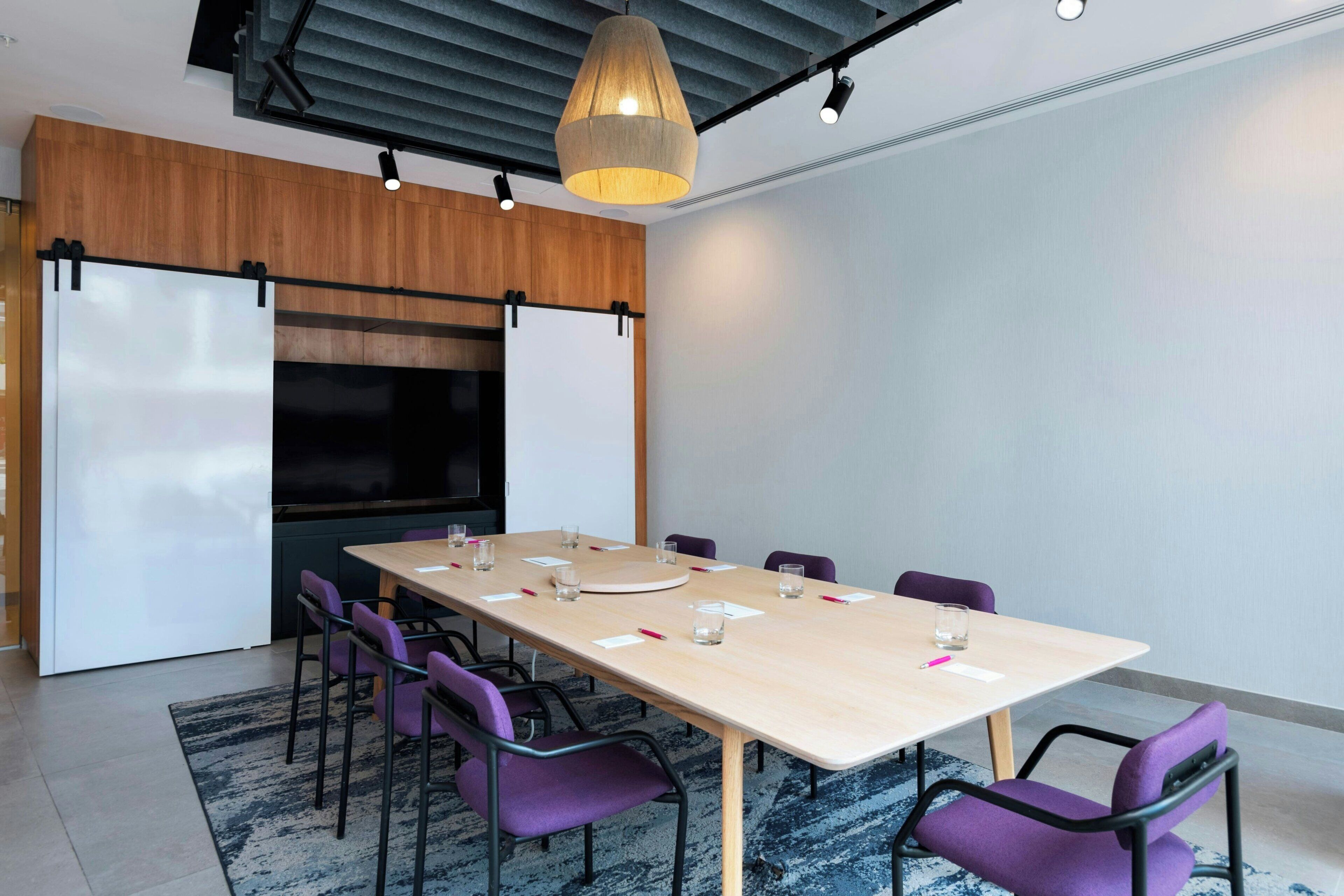 A conference room with a long table, purple chairs, a large screen, and a hanging light fixture.