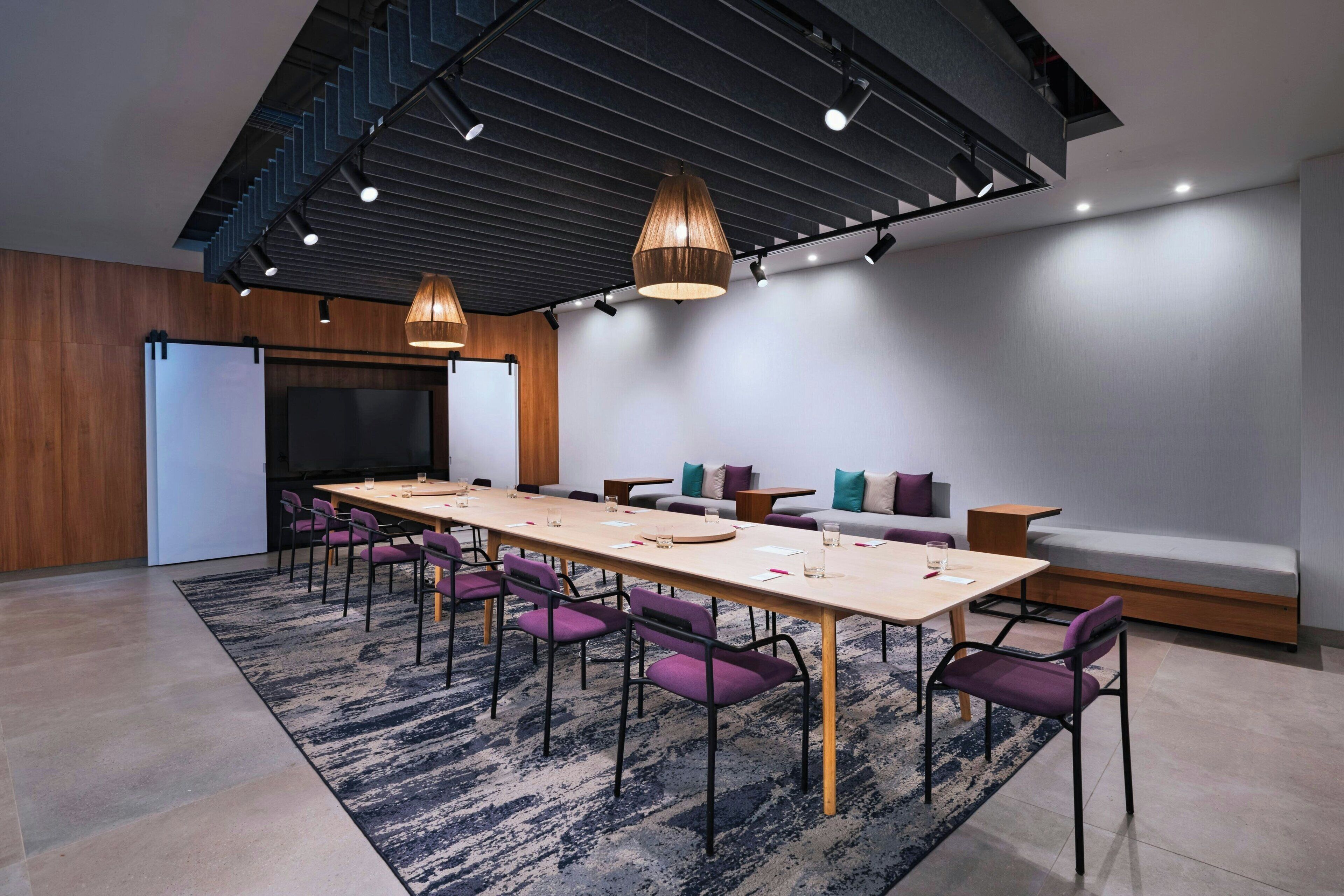 Una sala de reuniones moderna con una larga mesa, sillas moradas, un sofá e iluminación colgante.