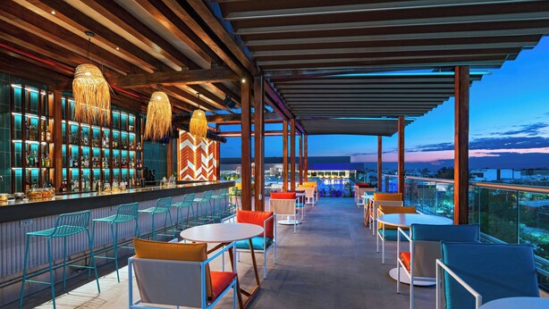 Un bar en la azotea con vistas al horizonte urbano, que incluye una zona de bar con estantes llenos de botellas, varias áreas de estar con sillas y mesas, y lámparas colgantes.