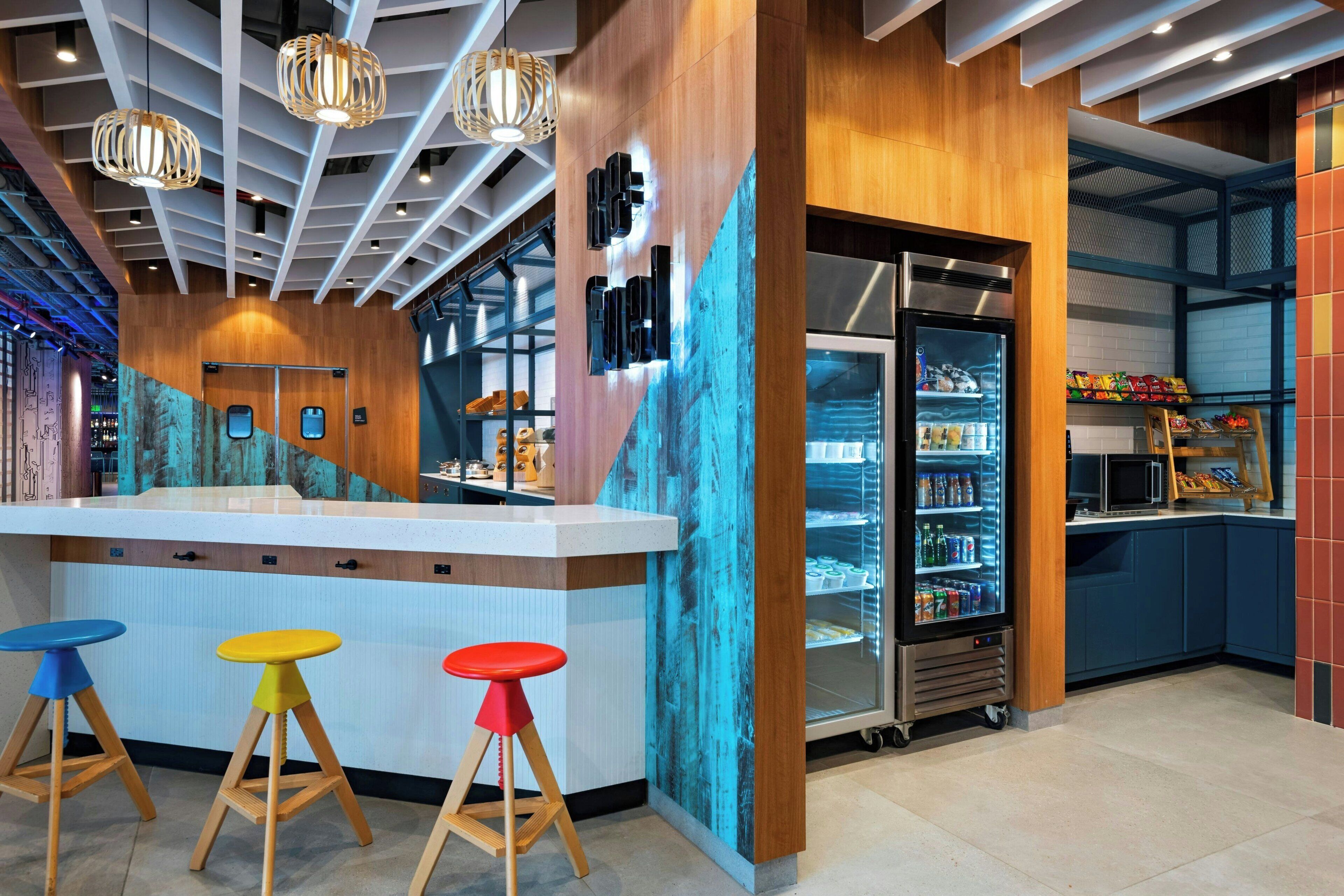 A modern bar area with a wooden counter, colorful stools, a refrigerated display case, and a blue mural on the wall.