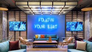 Un área de lounge moderna con un letrero de neón que dice "FOLLOW YOUR BEAT", dos pantallas grandes mostrando imágenes abstractas, un sofá turquesa, una mesa de centro de madera y sillas de mimbre.