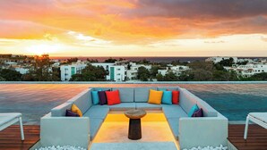 Zona de piscina en la azotea con mobiliario y vista a la ciudad al atardecer.