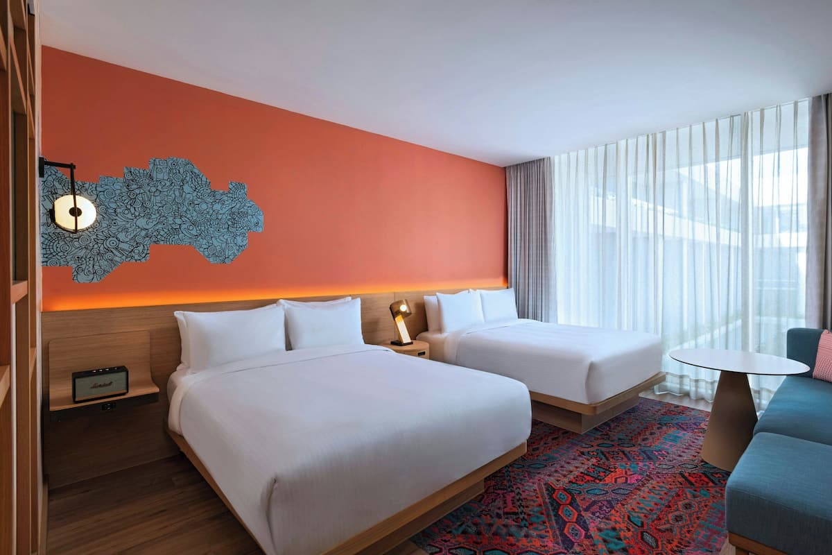 Une chambre d’hôtel avec deux lits, une petite table, un canapé, un élément décoratif mural et une grande fenêtre avec des rideaux.