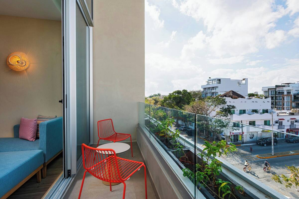 Un balcon avec un canapé et deux chaises rouges, donnant sur une rue bordée d’immeubles et fréquentée par des cyclistes.
