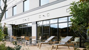 Un área de estar al aire libre con sillas y mesas de madera, ventanas amplias y fachada de edificio moderna.