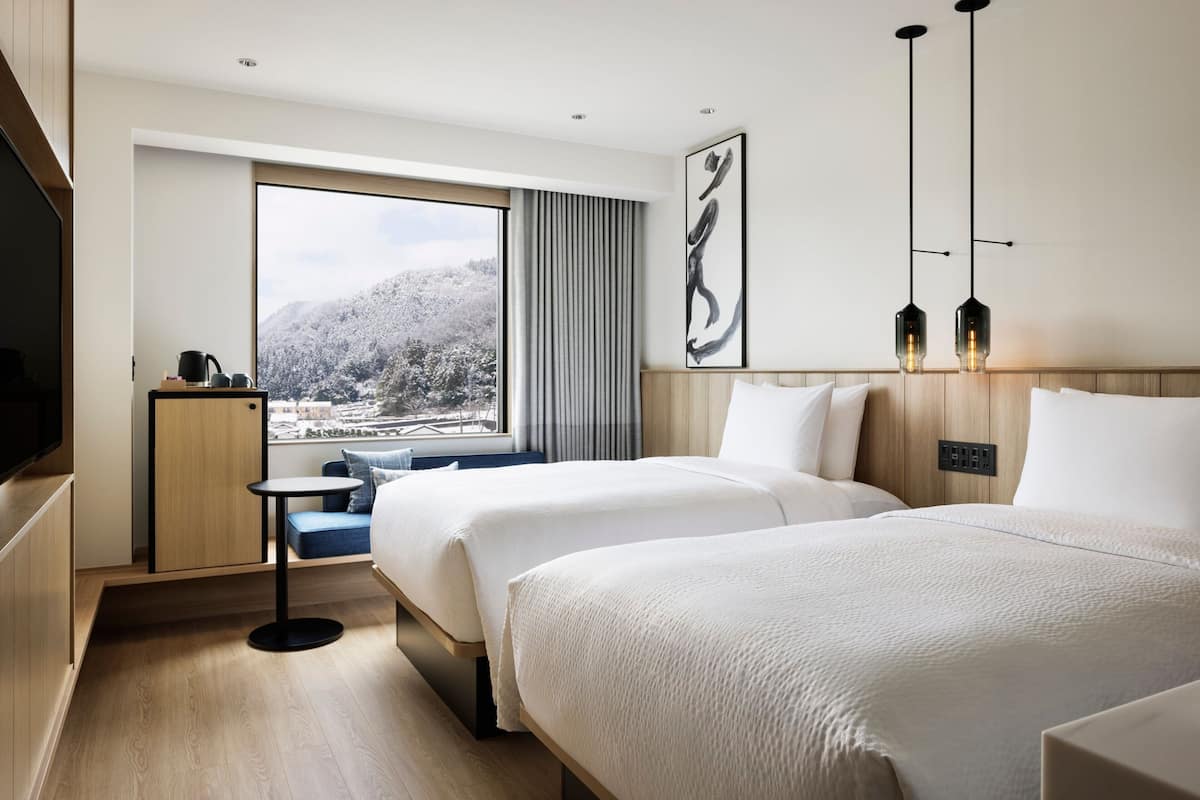 A modern hotel room with two beds, a small table, a TV, and a large window offering a view of a snowy landscape.
