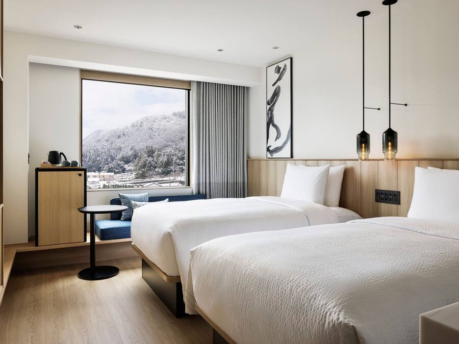 A modern hotel room with two beds, a small table, a TV, and a large window offering a view of a snowy landscape.