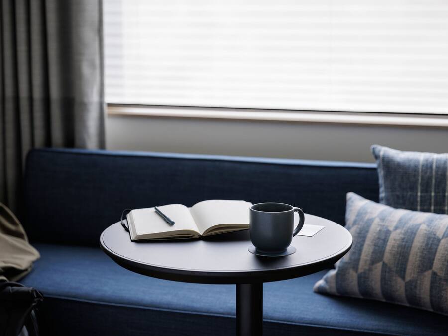 Una mesa redonda con un libro y una taza, sobre un sofá azul oscuro con un cojín estampado.