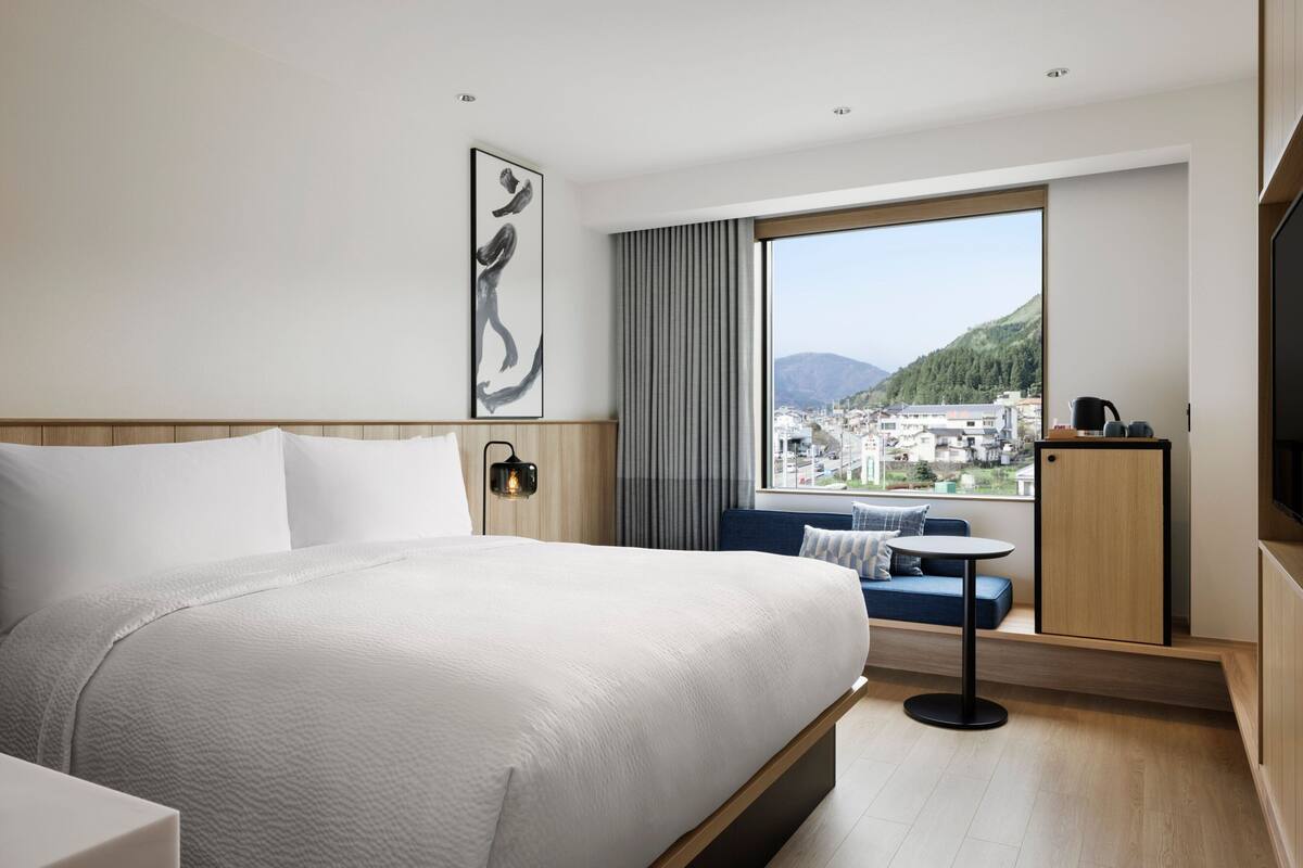 A modern hotel room with a large bed, a small table, a sofa, a TV, and a window with a city view.