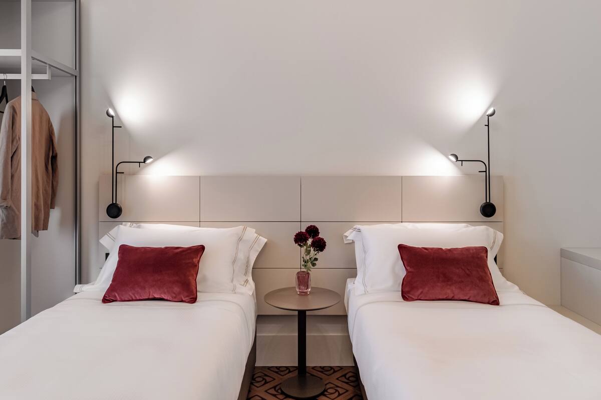 A hotel room with two beds, a small round table with a vase of flowers, and a wardrobe with hanging clothes.