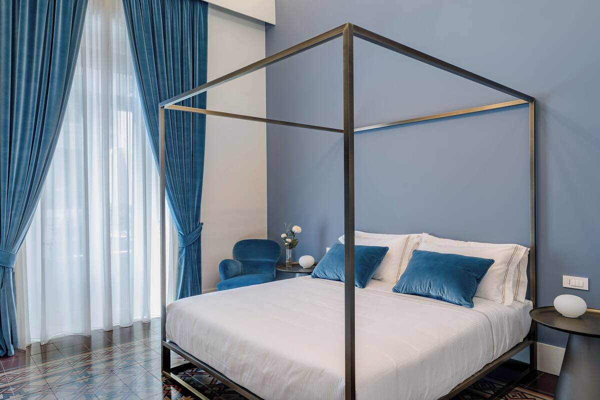 A four-poster bed with a white bedspread and blue pillows, a blue armchair, a small round table with a white vase, and a wall-mounted nightstand with a white lamp.