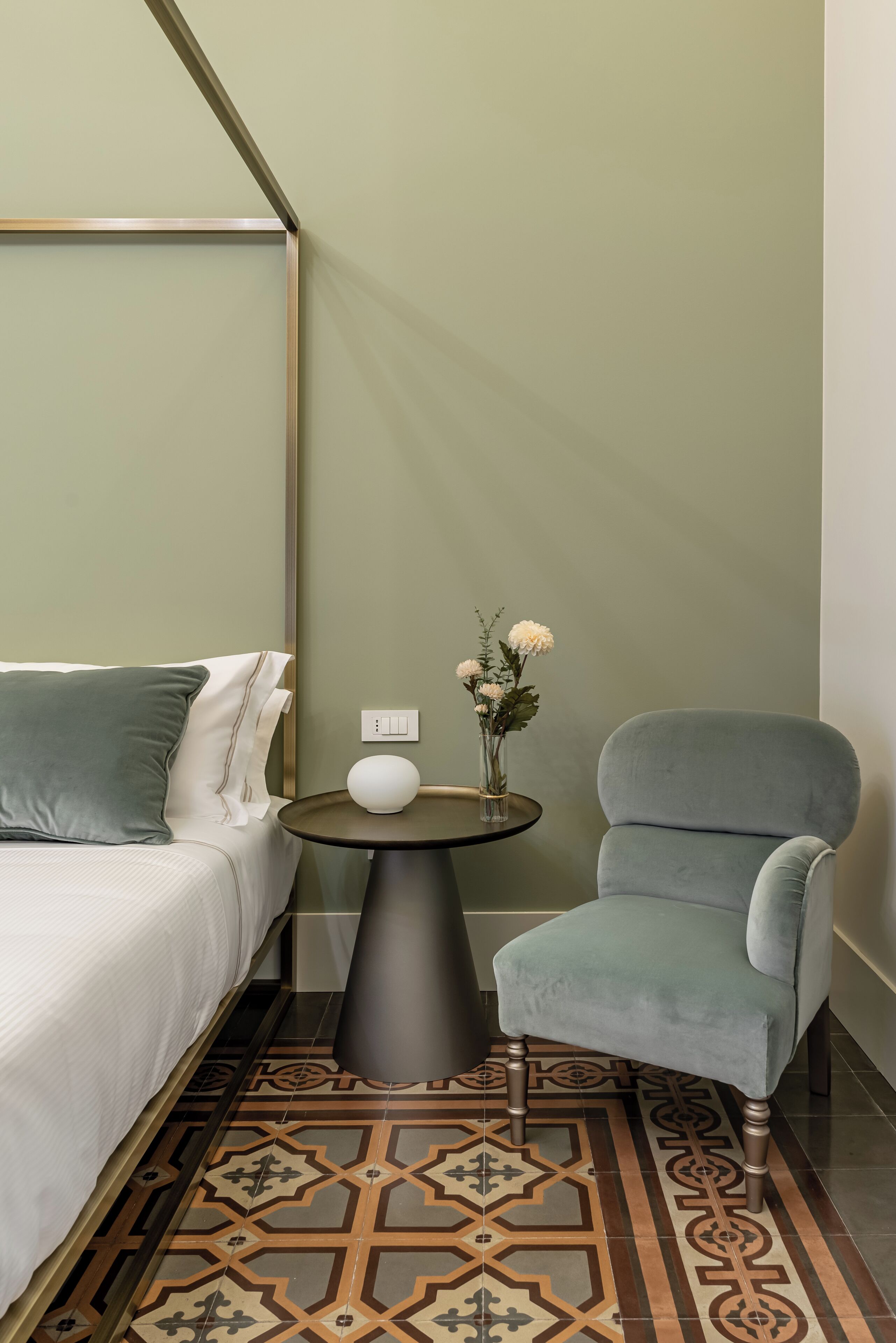 A hotel room with a bed, a chair, a small table with a vase of flowers, and a patterned rug.