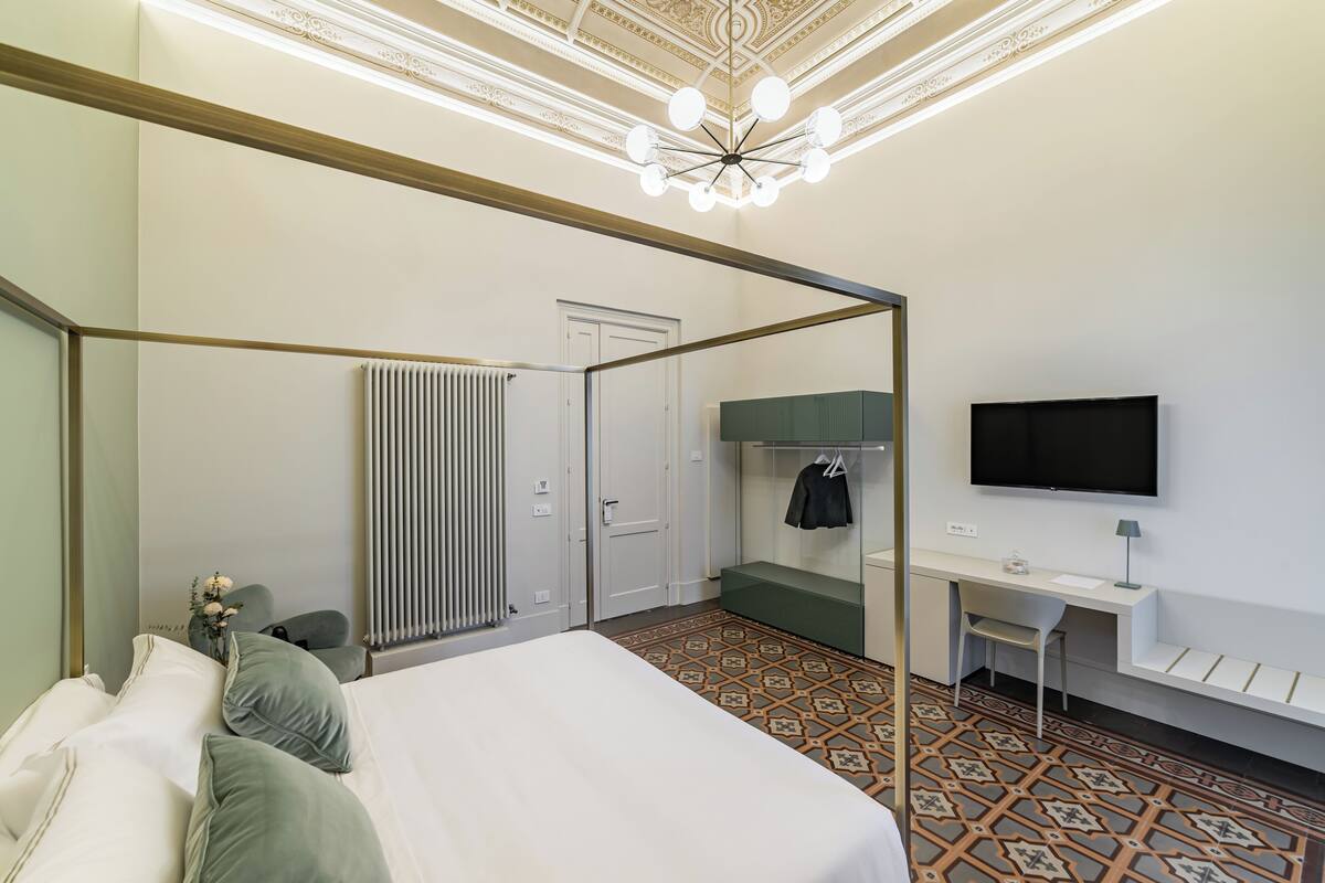 A hotel room with a four-poster bed, a desk with a chair, a television, and a radiator.