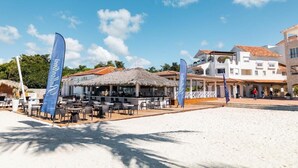 A beachside restaurant with outdoor seating, a thatched-roof structure, and a white building in the background.