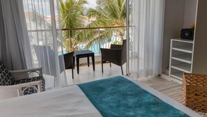 A balcony with a view of palm trees and a pool, furnished with a bed, a table, and two chairs.