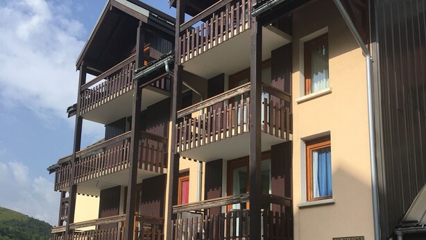 Een meerlaags gebouw met balkons, een bordje met de tekst 'LES BALCONS DE LONGCHAMP' en een parkeerplaats met een rode auto.